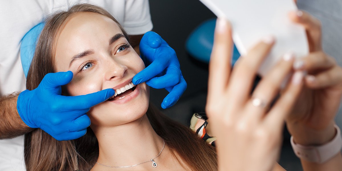 A woman smiling while a dentist in blue gloves checks her teeth with a mirror.