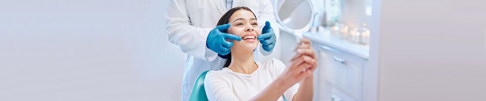 A smiling woman in a dental chair holding a mirror while the dentist shows her teeth.
