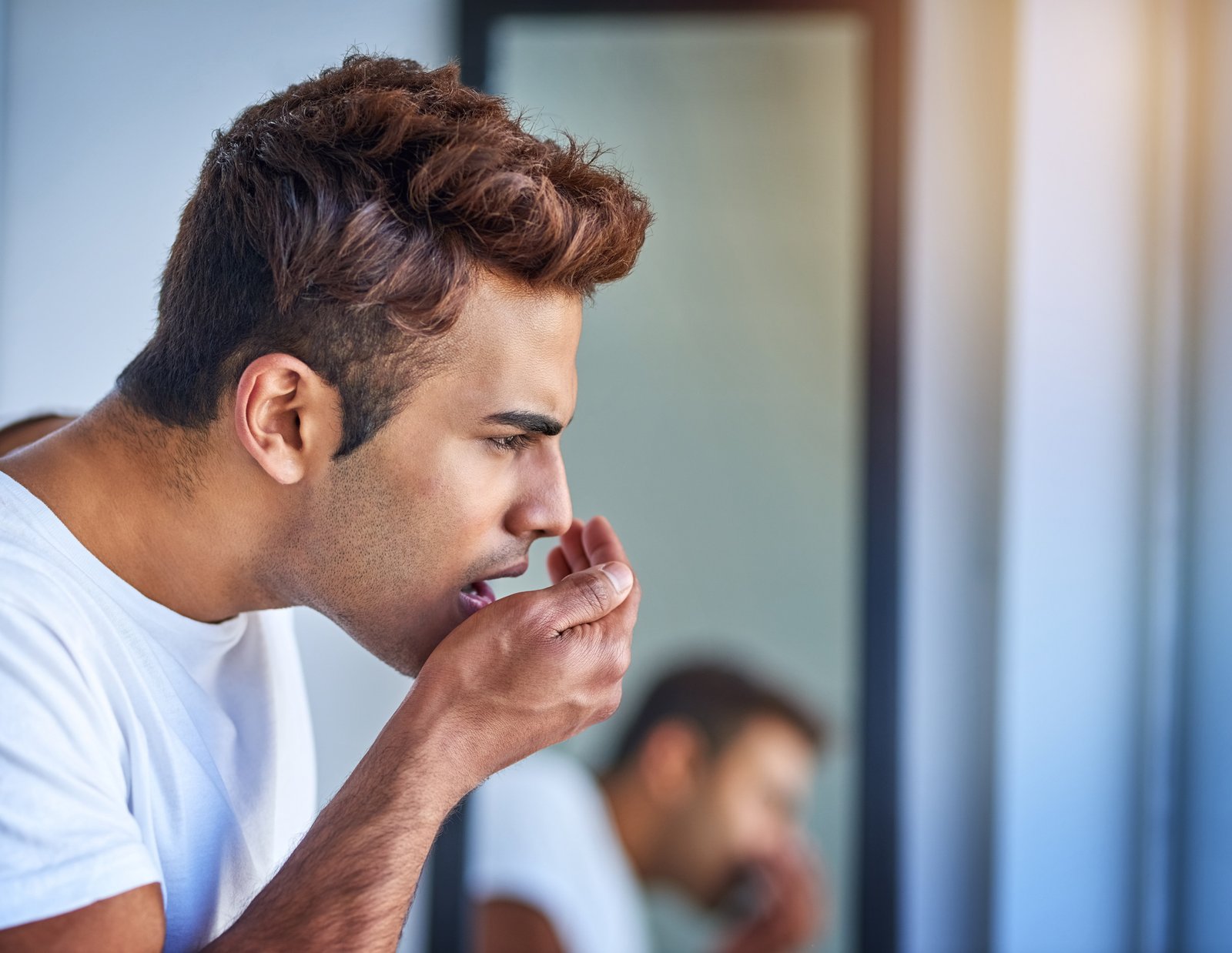 A man checking his breath by covering his mouth with his hand in front of a mirror.