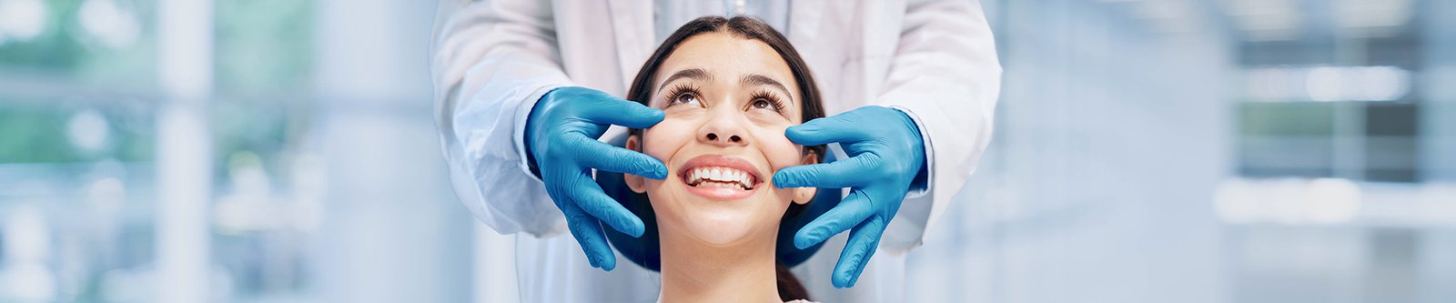 A smiling woman whose teeth are being examined by a dentist with blue medical gloves.