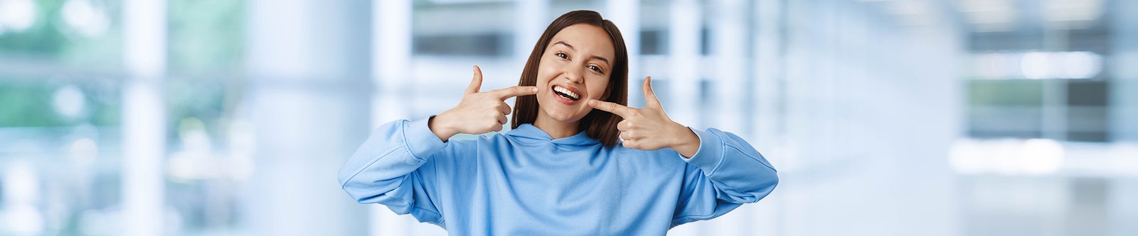 A smiling woman in a blue hoodie pointing to her white teeth.