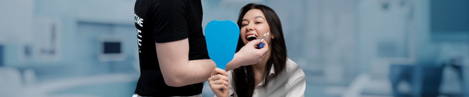 A woman holding a mirror while dentist matches veneer shades to her teeth.
